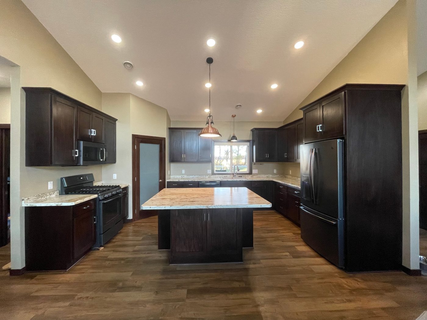 New construction kitchen in St. Cloud, MN with dark shaker cabinets, a large center island, laminate countertops, and a copper pendant light over the island under a vaulted ceiling